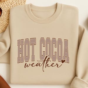 Peut inclure: Un sweat-shirt beige avec l'inscription "HOT COCOA weather" en marron. Le texte est dans une police de style vintage et universitaire. Le sweat-shirt a un col rond et des manches longues.