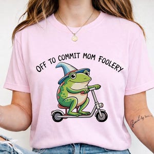 May include: A pale pink t-shirt with a cartoon frog on a scooter, wearing a wizard's hat. The frog is green with red spots. The text "OFF TO COMMIT MOM FOOLERY" is arched above the frog.
