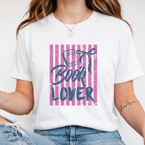 May include: White t-shirt with a pink and white striped design. The shirt displays a blue graphic of a book with a bow and the words "Book Lover" in a distressed font.