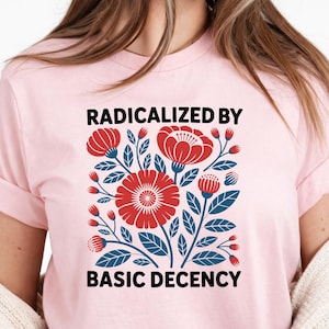 May include: Pale pink t-shirt with a floral design in red and blue, and the text "RADICALIZED BY BASIC DECENCY" in black. The shirt has short sleeves and a round neck.