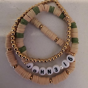May include: A set of three bracelets with gold beads and beige and green round beads. The bracelets are stacked on top of each other and spell out "JOHN 3 16".