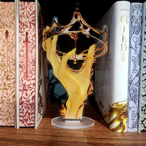 May include: A vertical acrylic standee featuring a stylised yellow hand holding a golden crown with a single droplet of liquid gold falling from the centre. The illustration is rendered in a glossy finish and is mounted on a clear circular base.