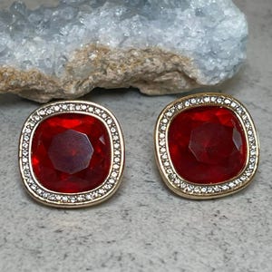 May include: A pair of square-shaped stud earrings. Each earring features a large, faceted, ruby-red gemstone set within a gold-toned frame. The frame is encrusted with small, clear crystals, adding sparkle. The earrings are displayed on a textured, neutral-toned surface.