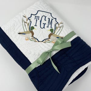 May include: A white and navy blue quilted baby blanket with a monogram of the letters "TGM" in a green frame with two embroidered ducks. The blanket is tied with a sage green ribbon.