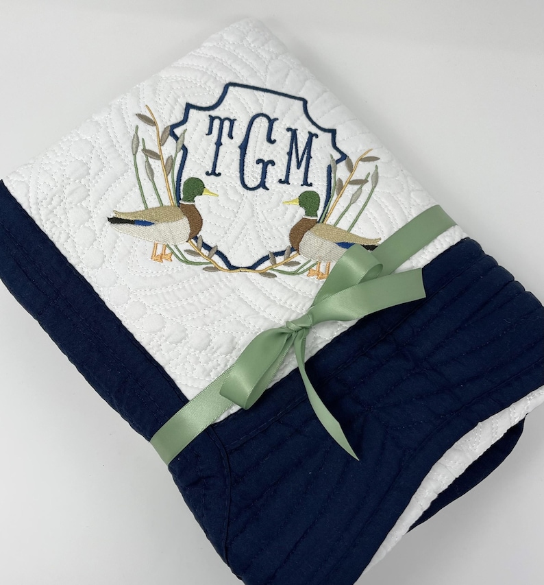May include: A white and navy blue quilted baby blanket with a monogram of the letters "TGM" in a green frame with two embroidered ducks. The blanket is tied with a sage green ribbon.
