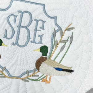 May include: A white quilted fabric with a blue embroidered monogram "SBE" and two embroidered mallard ducks on a branch with green leaves.