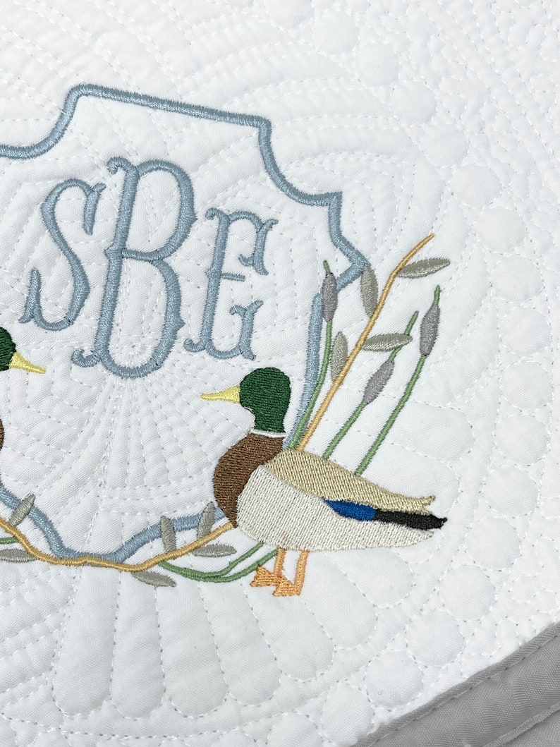 May include: A white quilted fabric with a blue embroidered monogram "SBE" and two embroidered mallard ducks on a branch with green leaves.
