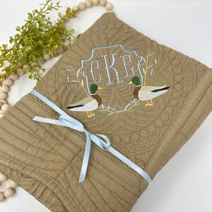 May include: A brown quilted throw blanket with a blue ribbon tied around it. The blanket features an embroidered design of two ducks and the monogram "CKE".