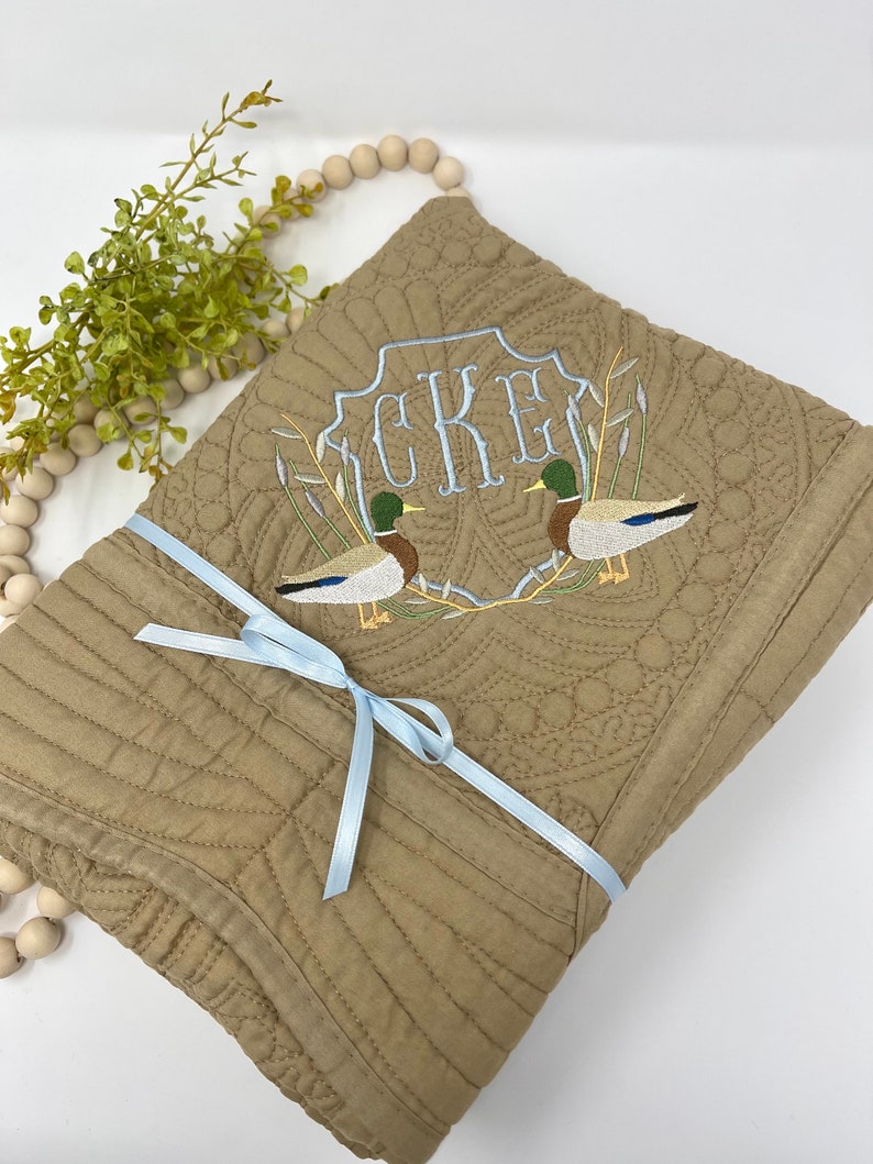 May include: A brown quilted throw blanket with a blue ribbon tied around it. The blanket features an embroidered design of two ducks and the monogram "CKE".