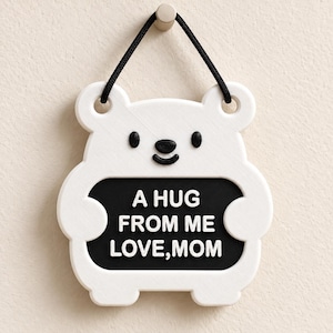May include: A white bear-shaped wall hanging with black eyes, a black nose, and a black mouth. The bear holds a black rectangular sign with the white embossed text A HUG FROM ME LOVE, MOM. A black cord is attached to the top for hanging purposes.