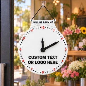 May include: A white circular sign with a black hanging cord, featuring the text WILL BE BACK AT above a clock face with red numbers and black hands. The center of the sign displays the black text CUSTOM TEXT OR LOGO HERE in bold letters.