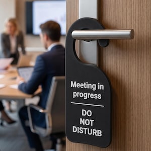 May include: A black door hanger with a textured surface hanging from a silver metal door handle. The sign features white raised text that reads Meeting in progress and DO NOT DISTURB, separated by a thin horizontal white line.