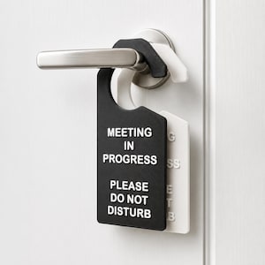 May include: A black door hanger with white raised text that reads MEETING IN PROGRESS PLEASE DO NOT DISTURB, layered over a white door hanger with matching text, both hanging from a brushed silver metal door handle on a white door.