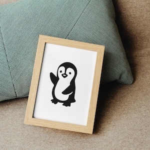 May include: A framed print featuring a black and white cartoon penguin waving. The penguin is centered on a white background within a light wood frame. The frame rests on a neutral-toned surface next to a teal pillow.