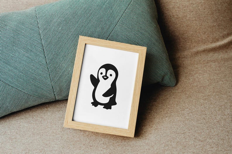 May include: A framed print featuring a black and white cartoon penguin waving. The penguin is centered on a white background within a light wood frame. The frame rests on a neutral-toned surface next to a teal pillow.