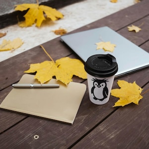 May include: A white coffee cup with a black lid and a penguin graphic sits on a wooden surface next to a closed laptop and a notepad with a pen. Yellow autumn leaves are scattered around.