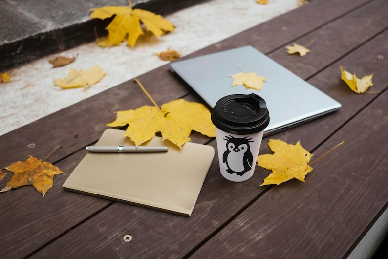 May include: A white coffee cup with a black lid and a penguin graphic sits on a wooden surface next to a closed laptop and a notepad with a pen. Yellow autumn leaves are scattered around.
