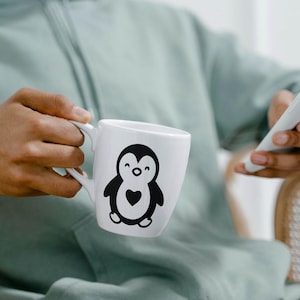 May include: White ceramic mug featuring a black and white penguin design with a heart. The mug is held by a person wearing a light green hoodie. A white smartphone is also visible.