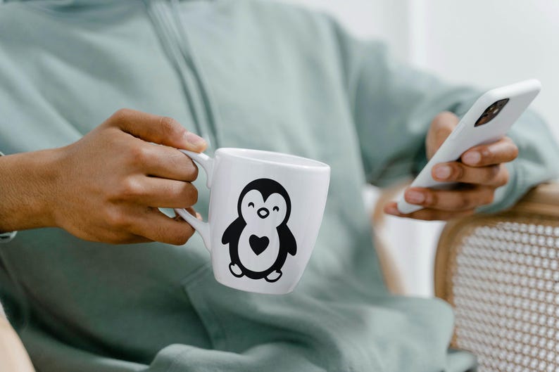 May include: White ceramic mug featuring a black and white penguin design with a heart. The mug is held by a person wearing a light green hoodie. A white smartphone is also visible.