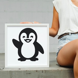 May include: A framed, black and white illustration of a cartoon penguin waving. The penguin has a white belly and a smiling face. The frame is white and square. The image is held by a person.