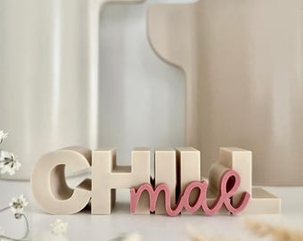 May include: A decorative sign spelling "CHILL mae" in a neutral color palette. The word "CHILL" is in block letters, while "mae" is in a cursive font. Two large, neutral-colored vases are in the background.
