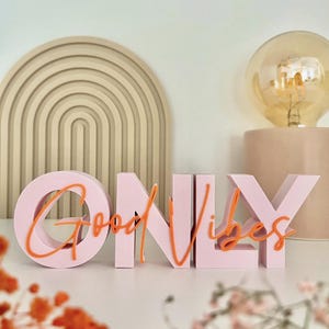 Op de afbeelding: Een roze decoratief bord met de tekst "ONLY" en de oranje tekst "Good Vibes". Het bord staat op een wit oppervlak, met een gloeilamp en een beige boogvormige wanddecoratie op de achtergrond.