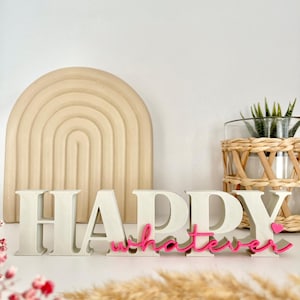 Könnte beinhalten: Ein dekoratives Schild mit dem Wort "HAPPY" in großen, cremefarbenen Blockbuchstaben und dem Wort "whatever" in rosa Schreibschrift. Ein kleines rosa Herz befindet sich neben dem Wort "whatever". Ein beigefarbener Bogen und eine Pflanze in einer Glasvase sind im Hintergrund.