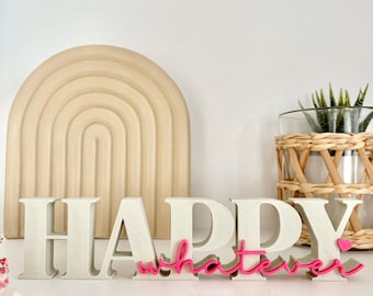 May include: A decorative sign with the word "HAPPY" in large, off-white block letters, and the word "whatever" in pink cursive. A small pink heart is next to the word "whatever". A beige arch and a plant in a glass vase are in the background.