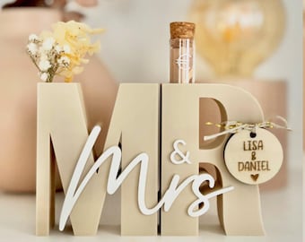 Puede incluir: Decoración de boda de madera beige con las letras "MR & Mrs" en fuente cursiva. Una pequeña etiqueta de madera con los nombres "Lisa & Daniel" cuelga de la "R". Un pequeño vial de vidrio con un tapón de corcho se encuentra encima. Flores secas se colocan en la "M".