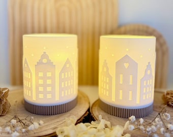 May include: Two cylindrical, off-white candle holders with a cut-out cityscape design. The candle holders sit on wooden slices and have a brown base. The background features a wooden arch and dried flowers, creating a cozy, decorative scene.