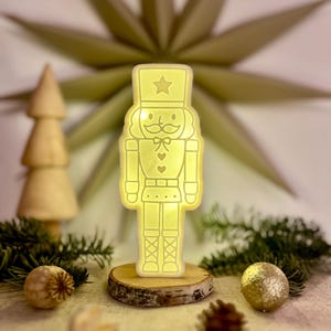 Może przedstawiać: Świecąca, żółta figurka LED dziadka do orzechów, stojąca na drewnianej podstawie. Figurka ma gwiazdę na kapeluszu i jest otoczona dekoracjami świątecznymi, w tym drewnianym drzewkiem, igłami sosnowymi i złotą błyszczącą ozdobą.