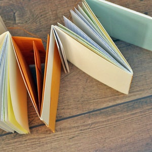 May include: A collection of handmade journals with different colored covers, including orange, blue, and white. The journals are open and show the pages inside, which are lined with different colors of paper.