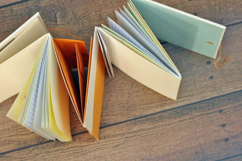 May include: A collection of handmade journals with different colored covers, including orange, blue, and white. The journals are open and show the pages inside, which are lined with different colors of paper.