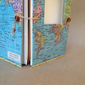 May include: A vintage-style world map journal with a blue background and colorful continents. The journal is closed and has a brown leather strap with metal buckles. The text "THE WORLD" and "MERCATOR PROJECTION" is printed on the front cover.