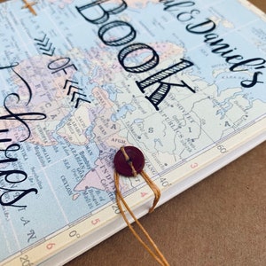 May include: A brown and white travel journal with a world map cover. The journal has a brown string closure and a brown button. The text "Abigail & Daniel's Book of the" is written on the cover.