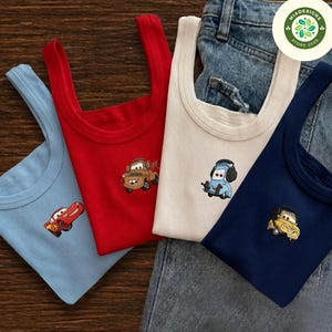 May include: Four ribbed tank tops in light blue, red, cream, and navy blue. Each tank top features a small embroidered character from the Cars movie franchise, including Lightning McQueen, Mater, Luigi, and Guido, centered on the chest area.