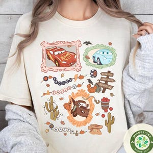 以下が含まれることがあります： クリーム色のTシャツ。カートゥーンの車のキャラクターが装飾フレームに入り、カボチャや葉など秋をテーマにしたイラストが描かれています。「KACHOW」と「SALLY」の文字入り。右下にストアロゴが見えます。