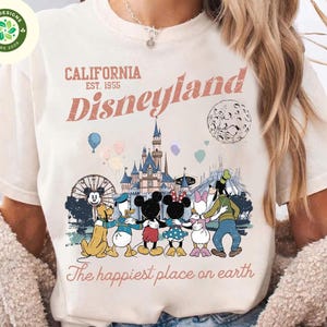 May include: Cream-colored t-shirt featuring a Disneyland graphic. The design includes the text "CALIFORNIA EST. 1955 Disneyland" above a colorful illustration of Disney characters and the castle. The phrase "The happiest place on earth" is printed below.