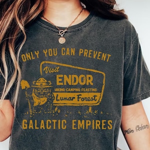 May include: A dark grey t-shirt with a vintage-style graphic design. The graphic features a cartoon character with a ranger hat and a sign that reads "Visit Endor Hiking-Camping-Feasting Lunar Forest". The text "Only You Can Prevent Galactic Empires" is printed above the graphic.