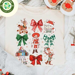 May include: Cream-colored crew neck t-shirt featuring a festive design with cartoon characters and bows. The design includes characters like Woody, Buzz Lightyear, and an alien, all adorned with Christmas lights and bows. The shirt is surrounded by holiday decor.