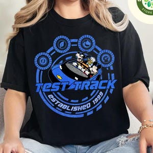 May include: Black t-shirt featuring a blue graphic of Mickey Mouse, Donald Duck, and Goofy in a race car, with the words "Test Track" and "Established 1999". The design is surrounded by a blue circular pattern.