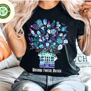 May include: Black t-shirt featuring a graphic of the Haunted Mansion from Disney World. The graphic depicts the mansion with ghosts and characters from the ride. The text "Welcome Foolish Mortals" is printed below the graphic.