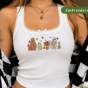 May include: A white ribbed tank top with a scoop neckline and lace trim. The center features a graphic print of four cartoon characters resembling Chewbacca, BB-8, an Ewok, and R2-D2, surrounded by autumn leaves and pumpkins. A small white bow is at the center.