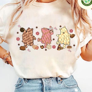 May include: Cream-colored sweatshirt featuring a whimsical illustration of three bear-like characters in patterned costumes. The bears are surrounded by decorative elements like stars, flowers, and food items. The design has a playful and cartoonish style.