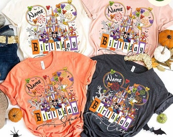 May include: Four short-sleeved t-shirts in cream, pink, orange, and charcoal gray, each featuring a colorful graphic. The graphic includes a castle, Mickey Mouse, and the word "Birthday". The shirts are hanging on a gold hanger.