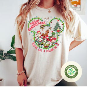 May include: Beige t-shirt featuring a festive design with the text "Merry Christmas" and "You've Got a Friend in Me." The design incorporates a Mickey Mouse head shape with Christmas lights, a Christmas tree, and Toy Story characters.