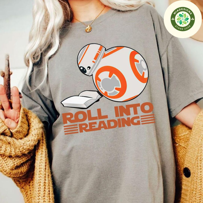 May include: A gray t-shirt with a white and orange robot character holding a book. The text "Roll into Reading" is printed in orange below the robot.