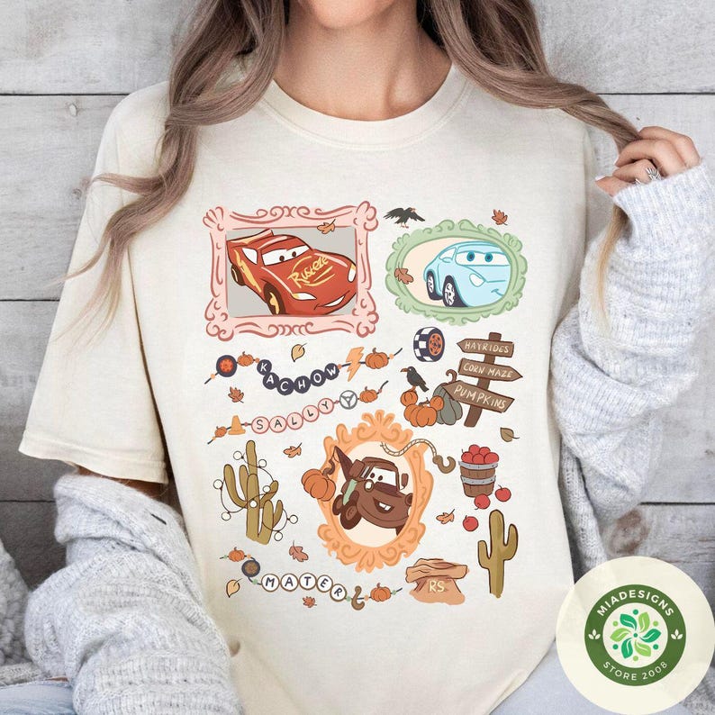 以下が含まれることがあります： クリーム色のTシャツ。カートゥーンの車のキャラクターが装飾フレームに入り、カボチャや葉など秋をテーマにしたイラストが描かれています。「KACHOW」と「SALLY」の文字入り。右下にストアロゴが見えます。