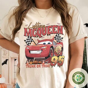 May include: Beige t-shirt featuring a graphic of Lightning McQueen from the movie Cars, with the text "Lightning McQueen" and "95". The design includes checkered flags, pumpkins, and the phrase "Trunk or Treat".