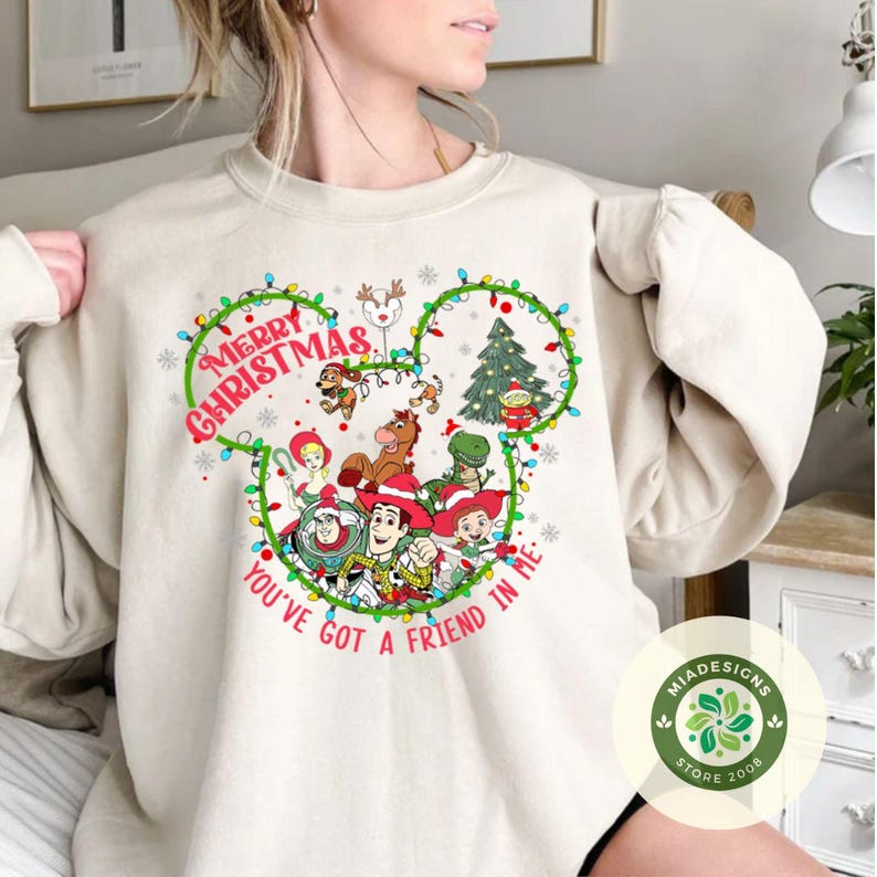 May include: Beige sweatshirt with a festive design featuring characters from a popular animated film. The design includes the text "Merry Christmas" and "You've Got a Friend in Me" in green, along with a Christmas tree and other holiday elements.